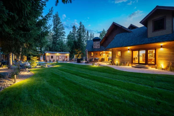 Expansive green lawn and landscaped backyard of a rustic-style home illuminated with warm outdoor lighting at dusk, surrounded by tall pine trees