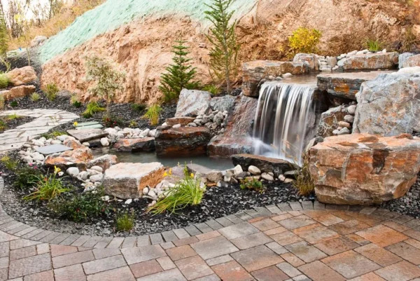 A beautifully landscaped backyard with a stone waterfall cascading into a small pond, surrounded by rocks, plants, and a brick pathway.