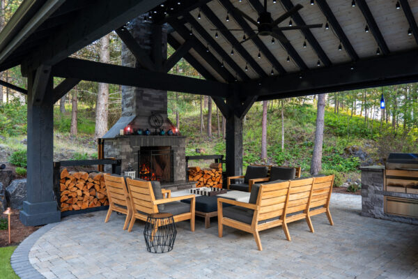 A covered outdoor living area with a stone fireplace, comfortable wooden furniture, and string lights overhead.